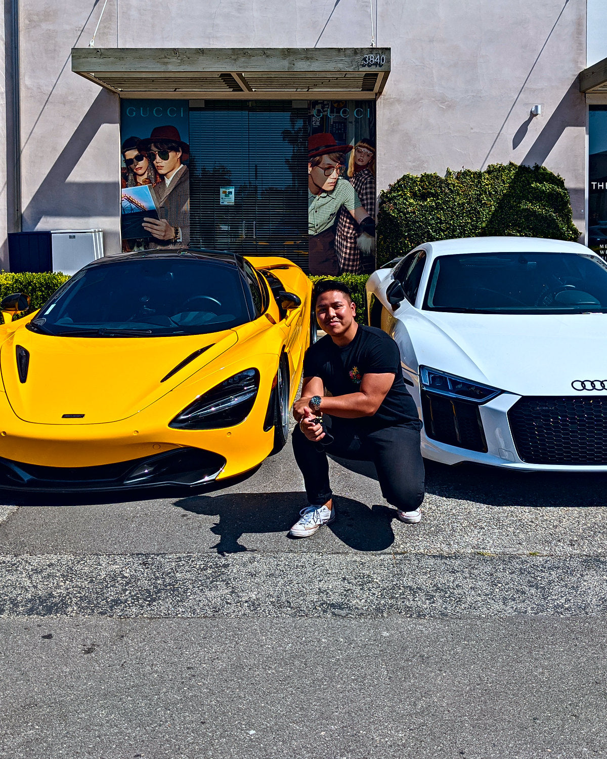 An owner crouched beside their yellow mid-engine supercar, a white mid-engine coupe parked next to it outside a storefront