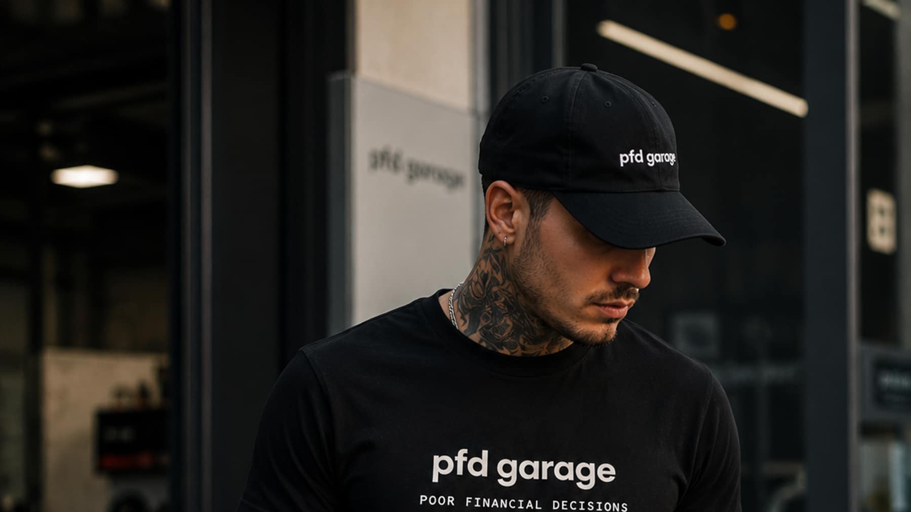 Man wearing a black cap and black tee, both printed with the pfd garage wordmark and a black, red and yellow bar, outside a garage storefront