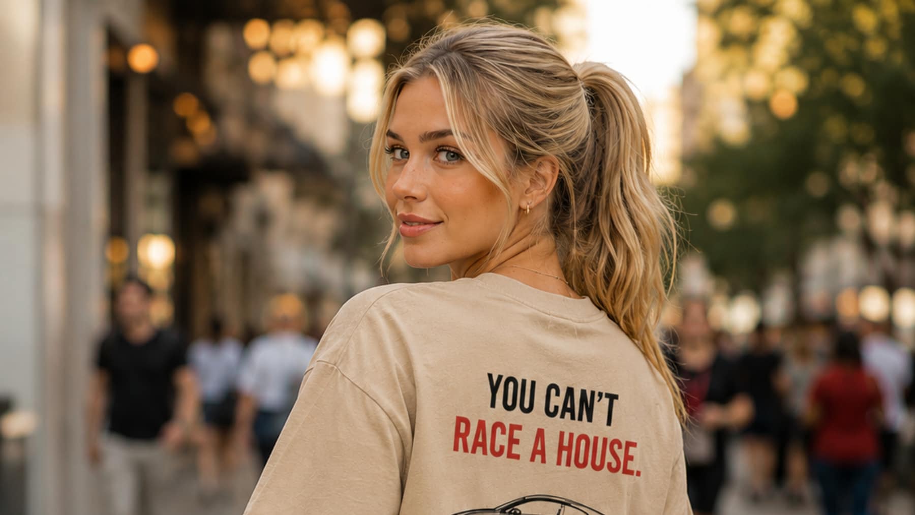 Back view of a sand tee printed You Can't Race A House above a line-drawing coupe and the pfd garage wordmark, worn on a city street at golden hour
