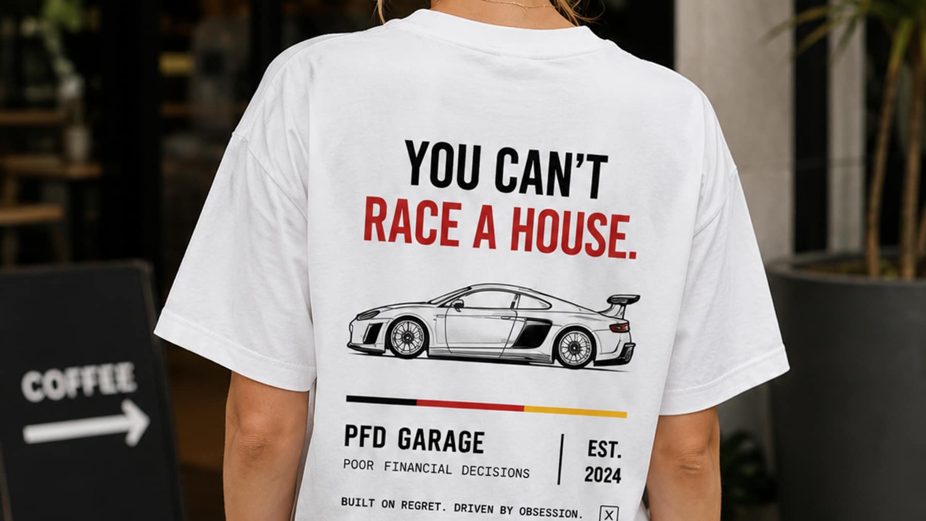Back view of a white tee printed You Can't Race A House above a line-drawing coupe and the pfd garage wordmark, worn outside a coffee shop