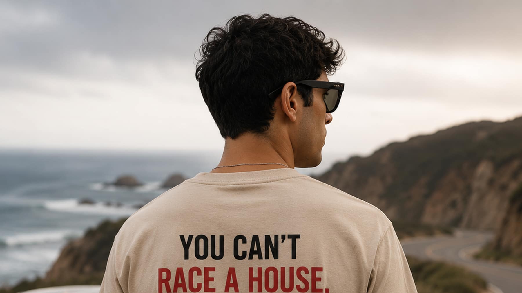 Back view of a sand tee printed You Can't Race A House above a line-drawing coupe, worn on a coastal road overlooking the ocean