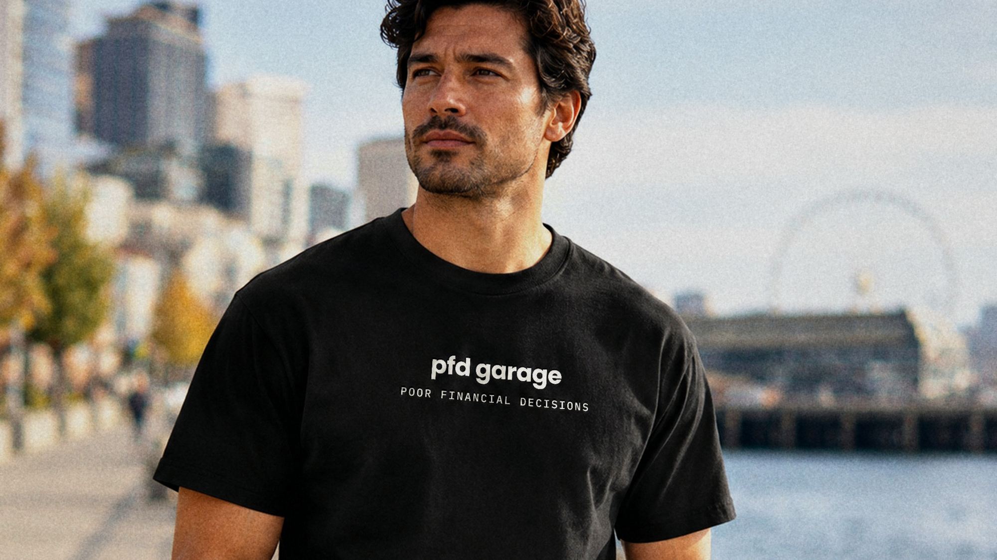 Man in a black tee with the pfd garage wordmark and Poor Financial Decisions across the chest, on a harbor promenade