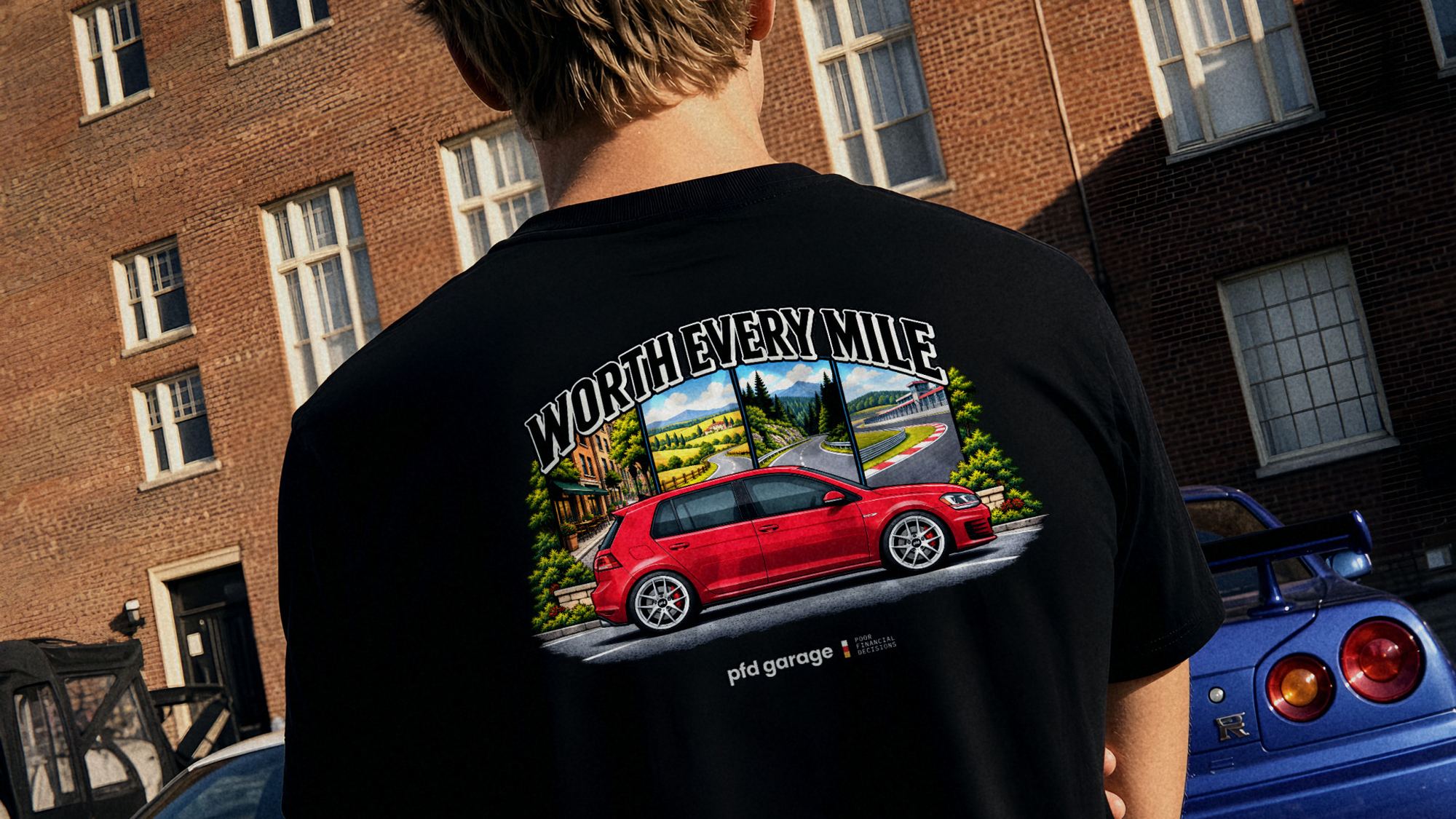 Back view of a man in a black Worth Every Mile tee with a red hatchback illustration, parked sports cars behind him