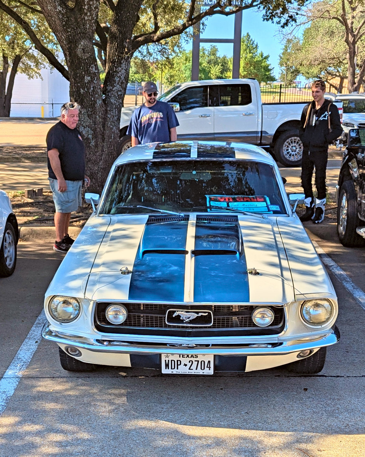 A white classic muscle coupe with blue stripes under oak trees at a meet
