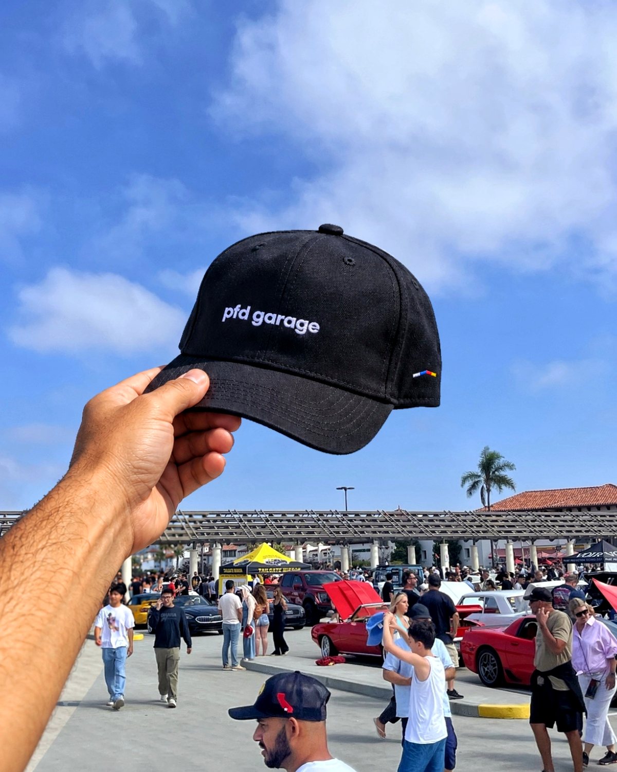 A black pfd garage cap held up against the sky at a busy car meet